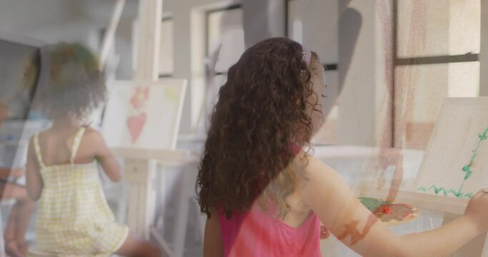 Animation of hand using paint brush over diverse schoolgirls painting at easels in art class
