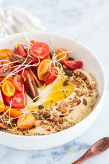 Hummus with tomatoes, seedlings, nuts and olive oil in white plate, white marble background.