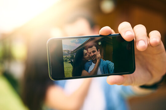 Happy couple, kiss and phone camera for selfie, photo or profile picture together in relationship outdoors. Hand of man holding smartphone with woman kissing cheek for love, memory or capture outside
