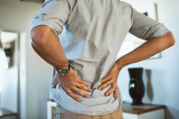 Hands, back pain and injury with a man standing in the living room of his home, holding his spine...