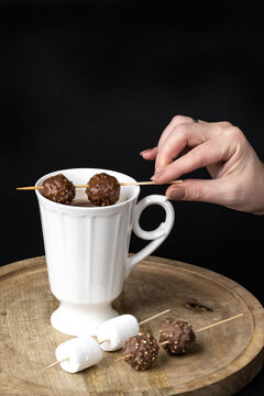 A woman's hand stirs hot chocolate with marshmallows and chocolates skewers in a beautiful white porcelain cup on a wooden table on a black background. Winter time. Festive concept.