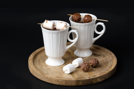 Two beautiful white porcelain cups of hot Chocolate drink with marshmallows and chocolates skewers over wooden table on black background. Winter time. Holiday concept..