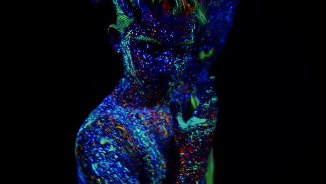A man painted with neon glowing colors poses on a dark background. Close-up of a man painted with fluorescent paints dancing with his hands, black background, front view