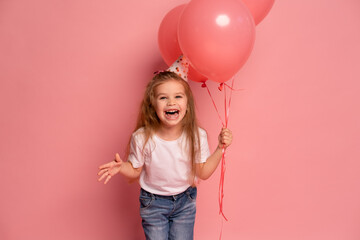little girl in birthday hat having fun, holding balloons on pink background