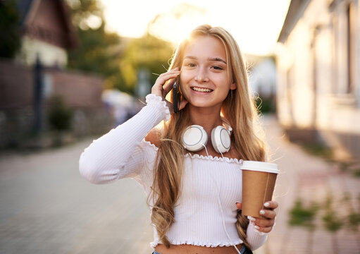 Young teeanager girl walking on a town street and talking on phone while holding paper coffee to go cup in her hand - Smiling student using her smartphone with headphones on her neck