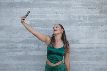 Latina and Hispanic girl, young and beautiful, recording a video with her cell phone, making funny gestures on a grey background. Concept mobile, smartphone, photo, video, apps, record.