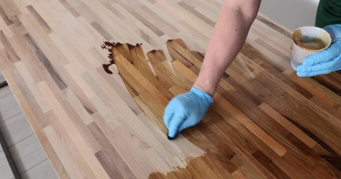 Worker in gloves polishes with varnish wooden floor surface standing at workplace in premise. Man uses liquid lacquer for processing wood slow motion