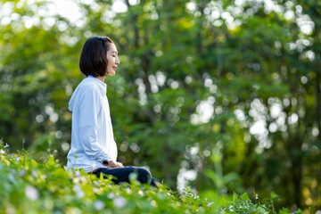 Asian woman is relaxingly practicing meditation yoga in forest full of wild flower in summer to...