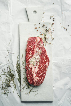 Raw marbled beef steak on white cutting board with herbs and salt. Top view