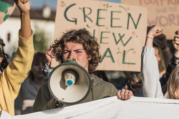 Young blond man with megaphone at environmental protest, "Green New Deal" sign visible.