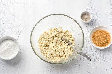 Glass bowl with mashed tofu, bowls with brown sugar, vanilla sugar and rice flour on a light blue background, top view. Cooking healthy vegan syrniki or other dishes
