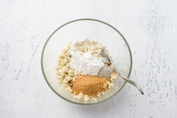 Glass bowl with mashed tofu, brown sugar, vanilla sugar and rice flour on a light blue background, top view. Cooking healthy vegan syrniki or other dishes