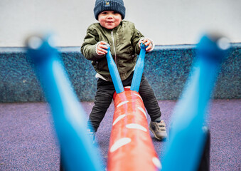 Happy child seen through the handles playing on a seesaw in a colourful playground. Playful kid outdoor on a dandle board conveys the importance of physical activity, recreation time and mental health - obrazy, fototapety, plakaty