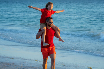 Son sits on fathers shoulders piggyback ride. Dad and son boy walking on beach. Summer holiday. Dad and son enjoying summer vacation together. Daddy and son playing on beach. Friendly family.