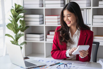 Asian businesswoman in red suit calculating company sales with calculator, laptop, and tablet on table Interior of businesswoman office at the modern workstation.