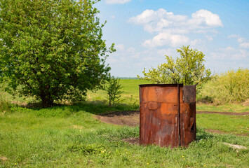 A large rusty garbage container stands in a clearing near trees on a spring day
