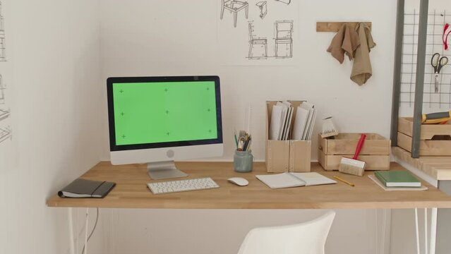 Arc shot of computer with green screen and wireless keyboard on desk in woodworking workshop with chair construction plans on wall, tools and files on workplace