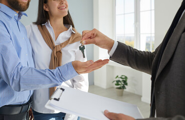 Realtor or real estate agent with a clipboard hands over the key to a young couple. Happy family buys a new house or apartment and takes the key from the realtor. Buying house concept - obrazy, fototapety, plakaty
