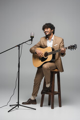 Smiling indian event host in formal wear playing acoustic guitar and singing in microphone in stand...