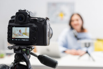 Digital camera on tripod standing near blurred happy female podcaster using devices and microphone on wooden table during stream in podcast studio