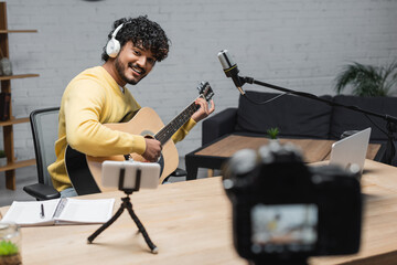 happy indian musician in headphones and yellow jumper playing acoustic guitar near smartphone on...