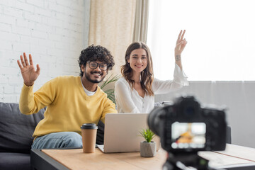 cheerful interracial vloggers in casual clothes waving hand and showing victory gesture during video call on laptop near blurred digital camera in radio studio