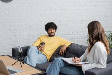 brunette woman writing in notebook near curly and smiling indian man in yellow jumper holding coffee to go while talking on couch near microphone and laptop in radio studio