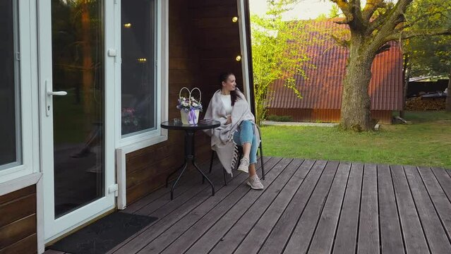 Cute woman wrapped in cozy blanket rests on veranda of tiny house