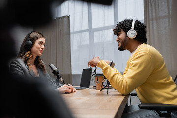 side view of smiling and curly indian man in headphones and yellow jumper talking to brunette...