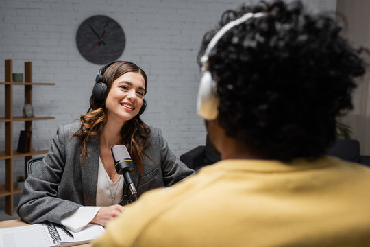 charming brunette broadcaster in headphones smiling near professional microphone, notebook and pen while talking to blurred indian man during interview in radio studio