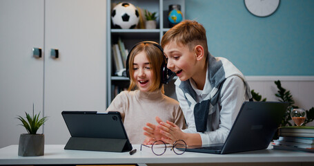 Small cute girl with headphones using internet games on digital tablet while kid boy playing game on laptop in living room at home, family concept