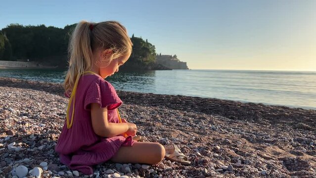 Little girl sits on the beach and collects pebbles in her purse