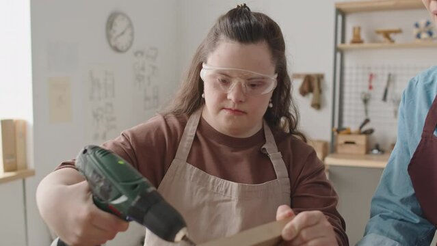 Tilt up shot of girl with Down syndrome in apron and protective eyewear using drill while making birdhouse with assistance of female teacher during woodworking workshop