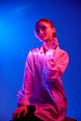 Tenderness. Portrait of young girl in white blouse with neon filter reflection posing against blue...