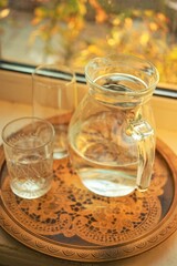 Jug of water and a glass in a wooden vintage tray on the windowsill.