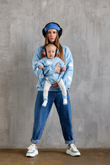 Full length portrait of a happy young mother and baby infant in her arms. Mom hugs her cute baby. Baby and mother in blue clothes and DJ headphones on their heads.