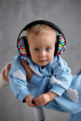 A happy young mother holds her precious baby in her arms. The kid looks into the camera with gray eyes and cheerful multi-colored DJ headphones on his head.