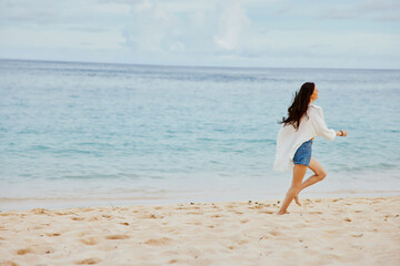 Naklejka premium Sports woman runs along the beach in summer clothes on the sand in a yellow T-shirt and denim shorts white shirt flying hair ocean view, beach vacation