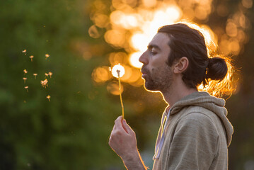 Funny man blowing dandelion over blured green grass, summer nature seeds fly outdoor light long hair Portrait of young bearded  golden sunset background make a wish.