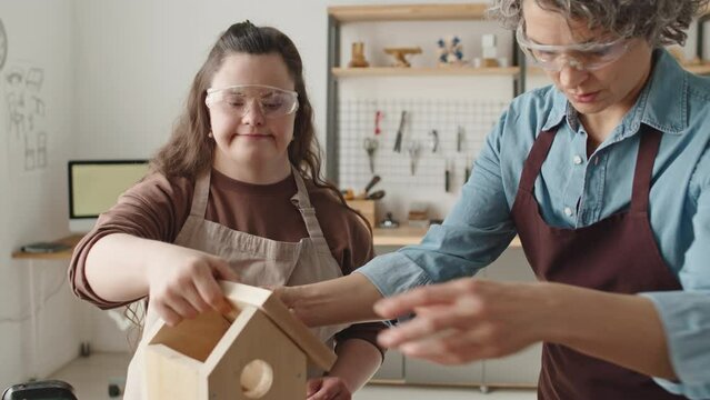 Girl with Down syndrome in apron and protective eyeglasses helping female teacher with assembling wooden birdhouse during masterclass in carpentry workshop