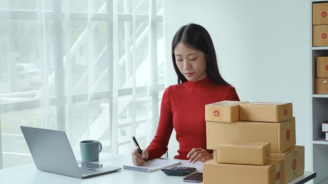 e-commerce, small business, sme , beautiful Asian woman looking at  note and using calculator to calculate taking customer orders and earning from online sales with smiling face happy while working