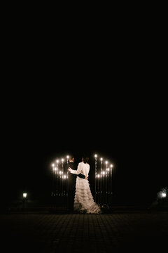 Young bride and groom stand near the wedding arch at night with lights, smoke and fireworks, silhouettes of the newlyweds