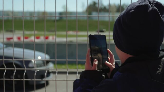 Little boy in hat and jacket takes pictures of sports car passing behind fence of racing track on phone. Young fan without seat on podium on sunny day shoots video for social networks.