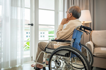 Asian senior man disabled sitting alone in wheelchair looking through window at hospital, lonely elder thoughtful sad old man look outside windows in bedroom at retirement home, feel depressed lonely
