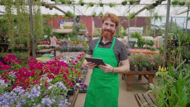 A male staff of Flower Shop holding tablet device while standing inside local business smiling at camera. Young redhair man employee wearing apron and using modern technology