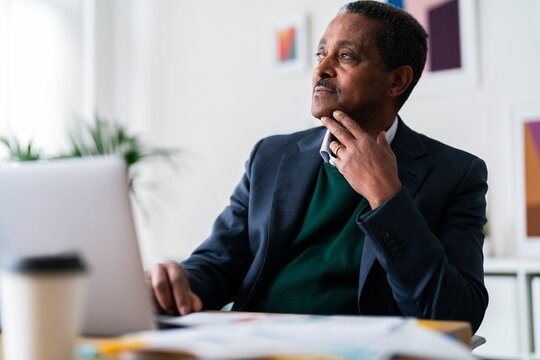 Focused elderly Ethiopian male CEO manager with laptop looking away and contemplating while creating business plan and working on project in workplace