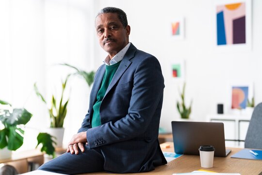Portrait of mature Ethiopian male executive manager sitting on desk near computer with coffee and looking at camera while working in spacious workplace
