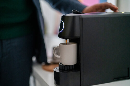 Selective focus of crop unrecognizable Ethiopian male entrepreneur with ring waiting near capsule coffee maker while filling mug with hot drink in modern office