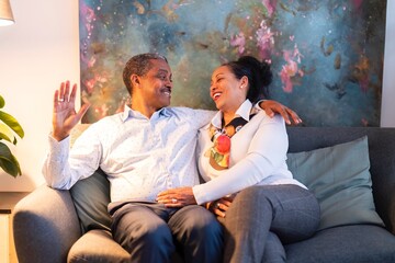 Positive adult Ethiopian married couple in elegant clothes sitting on sofa and hugging while smiling and looking at camera during romantic time together at home