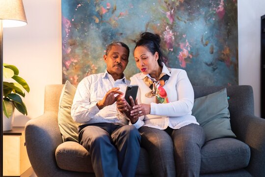Concentrated adult Ethiopian married couple in elegant clothes sitting on comfortable sofa and sharing smartphone together while surfing internet during weekend at home